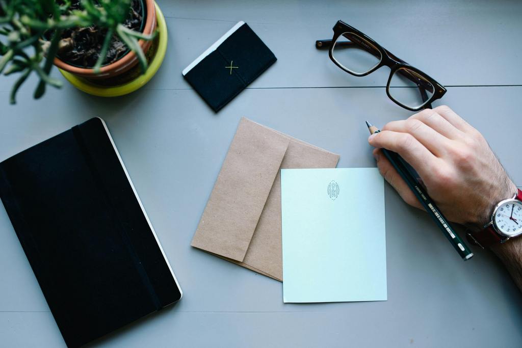 A person's hand holding a pencil, writing on a light blue card, with a stack of two brown envelopes, a black notebook, a small black wallet, glasses, and a potted plant on a gray surface. What’s the Best Way to Generate Leads Online? (Without Chasing People or Getting Ghosted)