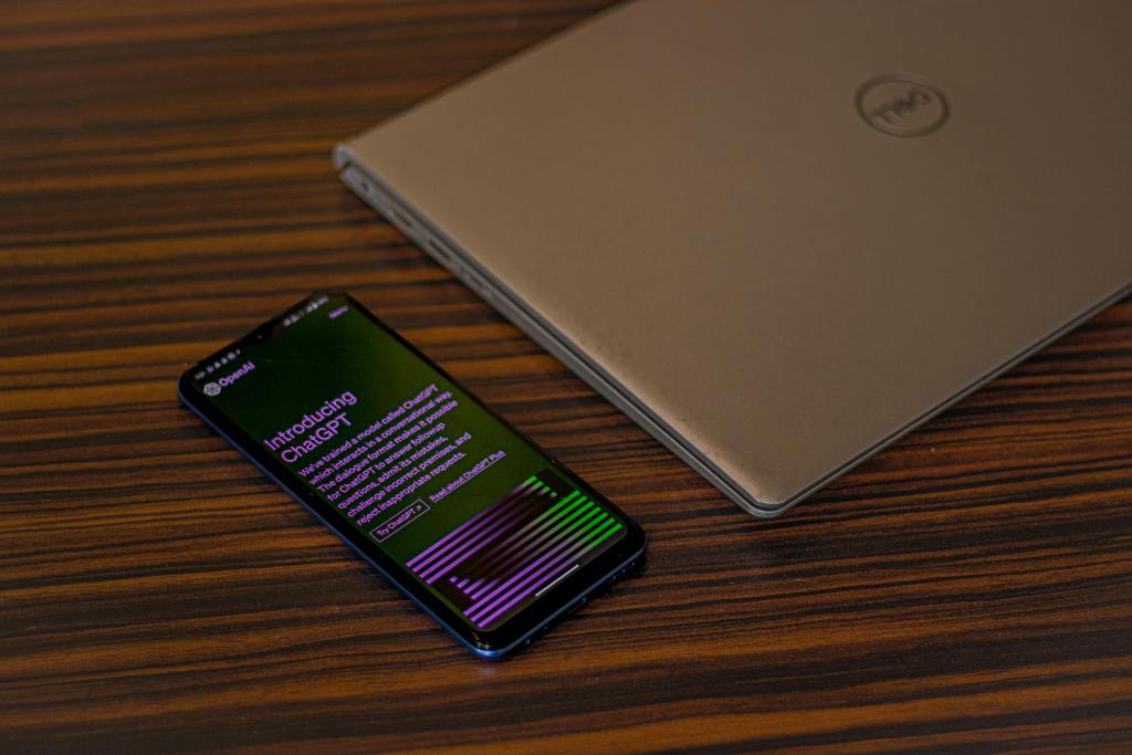 A smartphone displaying the 'Introducing ChatGPT' screen alongside a closed Dell laptop on a wooden table.